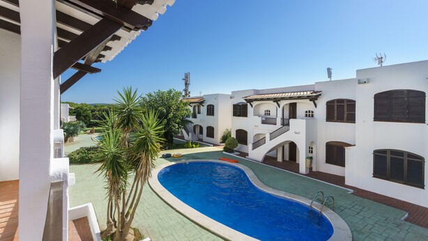 A swimming pool surrounded by a courtyard with a white building, a palm tree, and a tiled roof. - Apartamentos Posidonia (Mercadal)