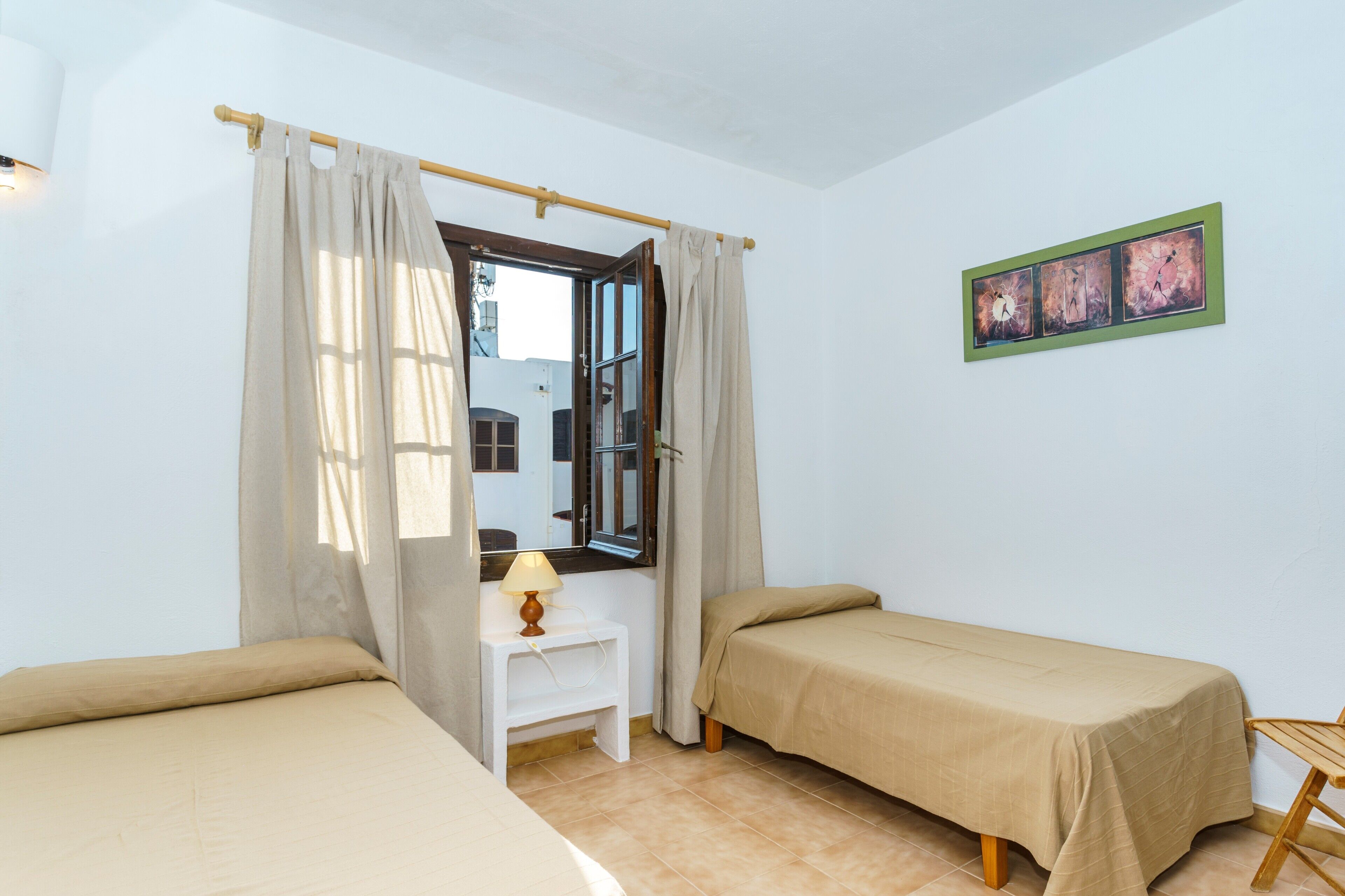 Une chambre avec deux lits, une petite table avec une lampe, une fenêtre avec des rideaux et un tableau accroché au mur.