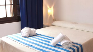 A hotel room with a bed, a nightstand, a wall-mounted lamp, and a window with curtains. - Apartamentos Posidonia (Mercadal)
