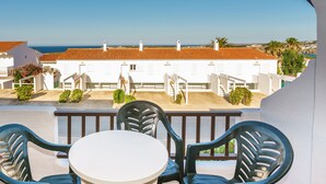 A balcony with a white table and two chairs, overlooking a white building with a red-tiled roof and a clear blue sky. - Apartamentos Posidonia (Mercadal)