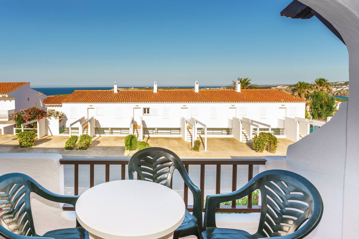 Un balcon avec une table et des chaises blanches, donnant sur une rangée de bâtiments blancs aux toits recouverts de tuiles rouges et offrant une vue dégagée sur la mer.