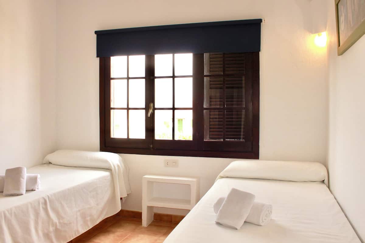 Une chambre avec deux lits simples, une fenêtre avec des stores, une petite table de chevet blanche et une lampe fixée au mur.