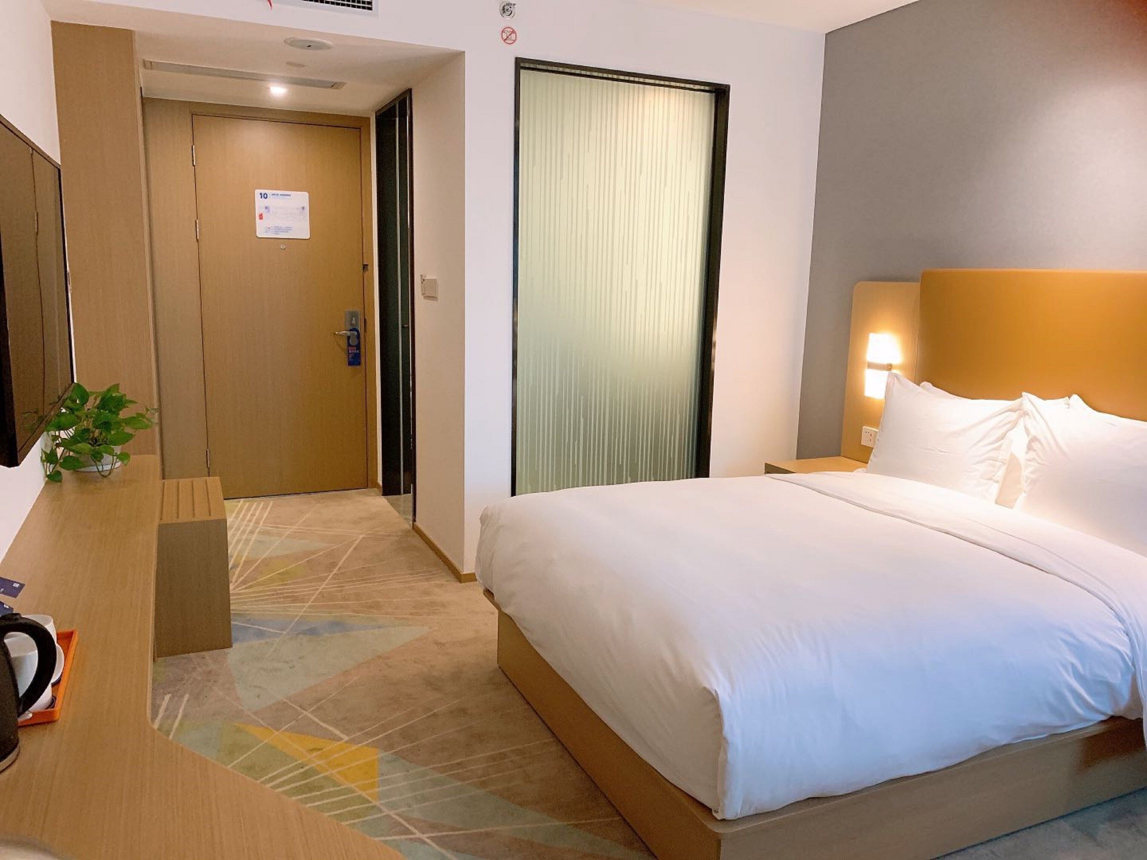 Une chambre d’hôtel avec un lit, un bureau agrémenté d’une plante, une porte et un luminaire fixé au mur.