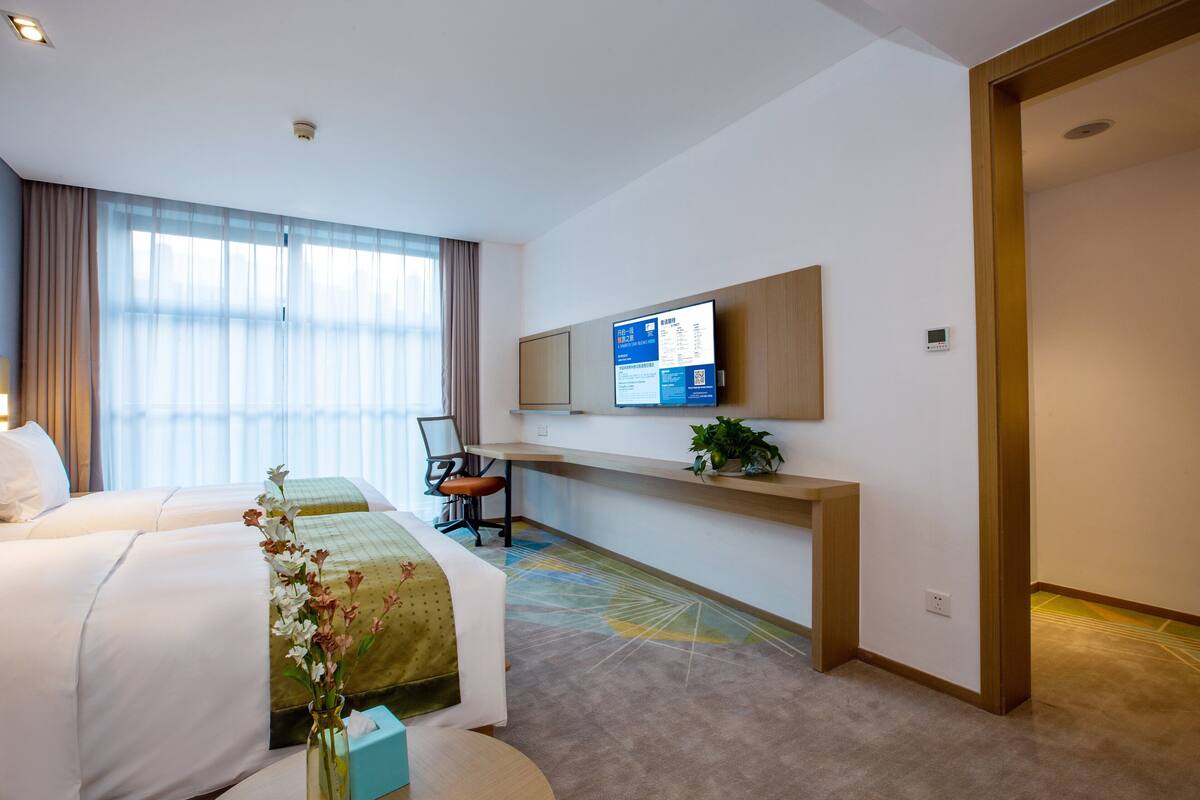 A hotel room with two beds, a desk, a chair, a TV, and a plant.
