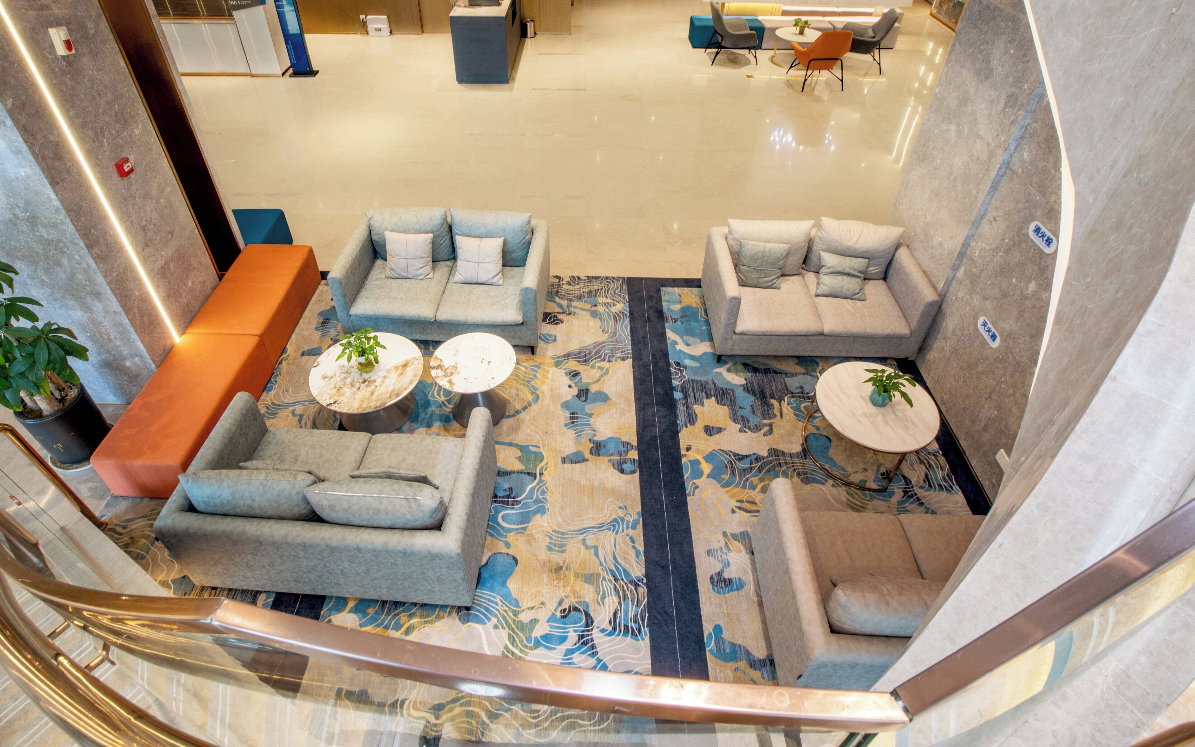 A modern hotel lobby with a spacious seating area featuring grey sofas and armchairs, a patterned carpet, and a staircase with a glass railing.