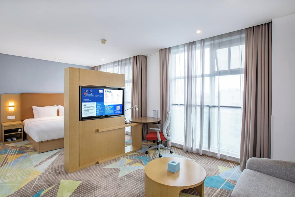 A hotel room with a bed, a desk with a computer, a chair, a small table, and a large window with curtains.