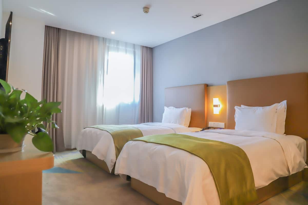 A hotel room with two beds, beige curtains, a potted plant, and a wall-mounted light fixture.