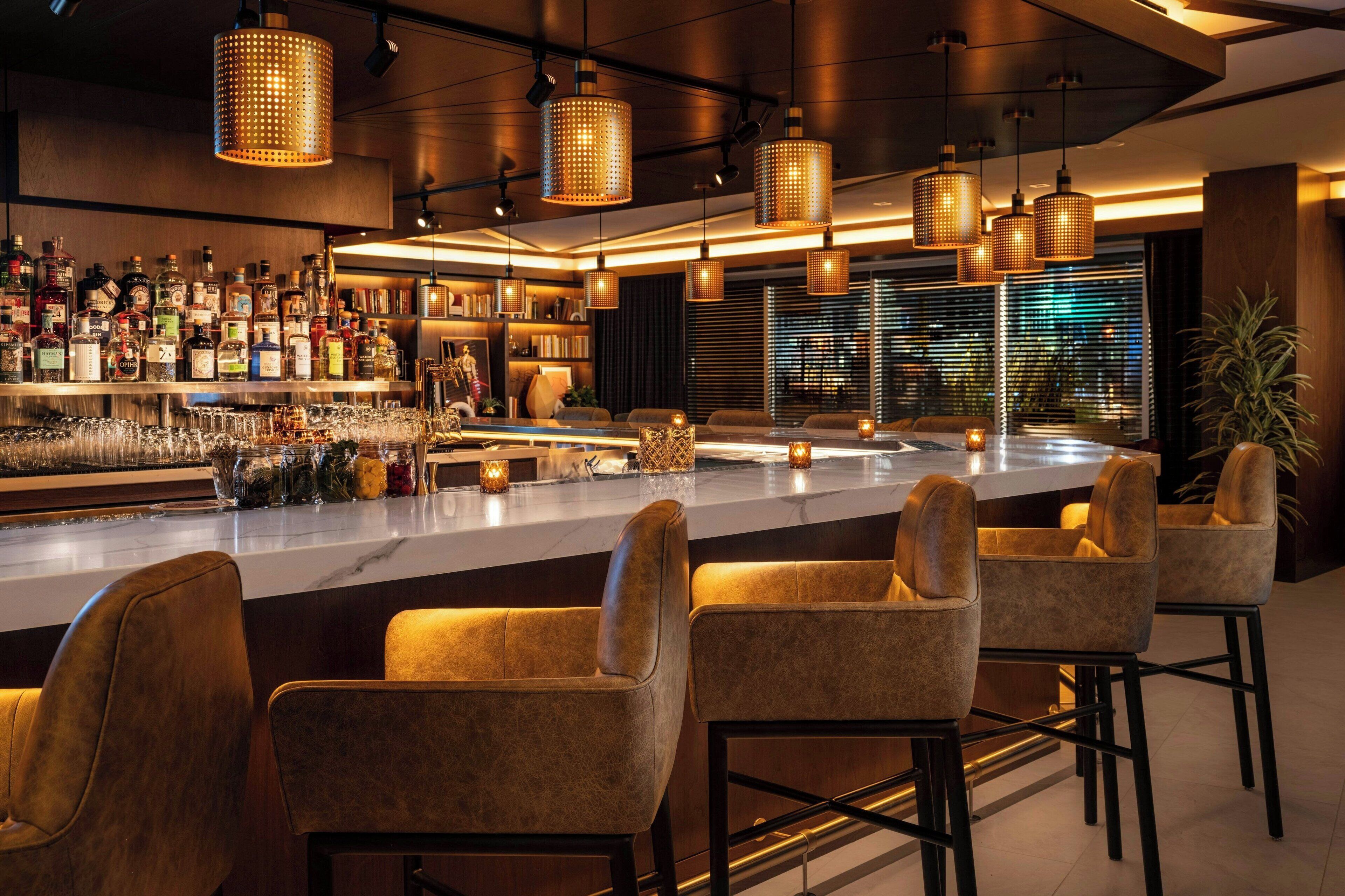 A bar area with a long counter, multiple bar stools, and hanging lights.