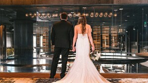 A couple standing in front of a reflective glass wall with a modern interior design, including a circular seating area and vertical light fixtures. - The Otis Hotel Austin, Autograph Collection (Austin)