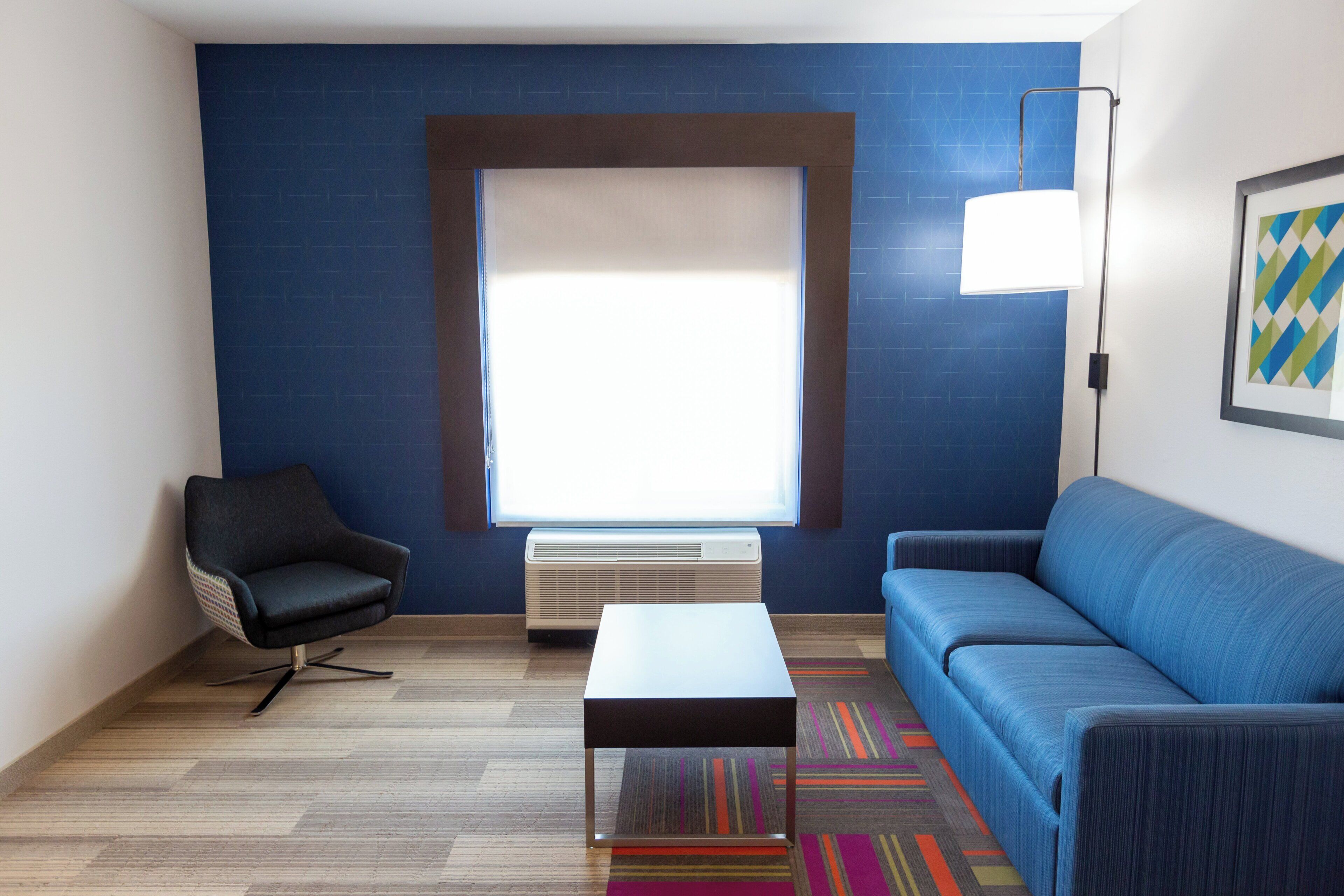 Een moderne hotelkamer met een blauwe accentmuur, een groot raam, een zwarte stoel, een blauwe bank, een witte salontafel en een kleurrijk vloerkleed.