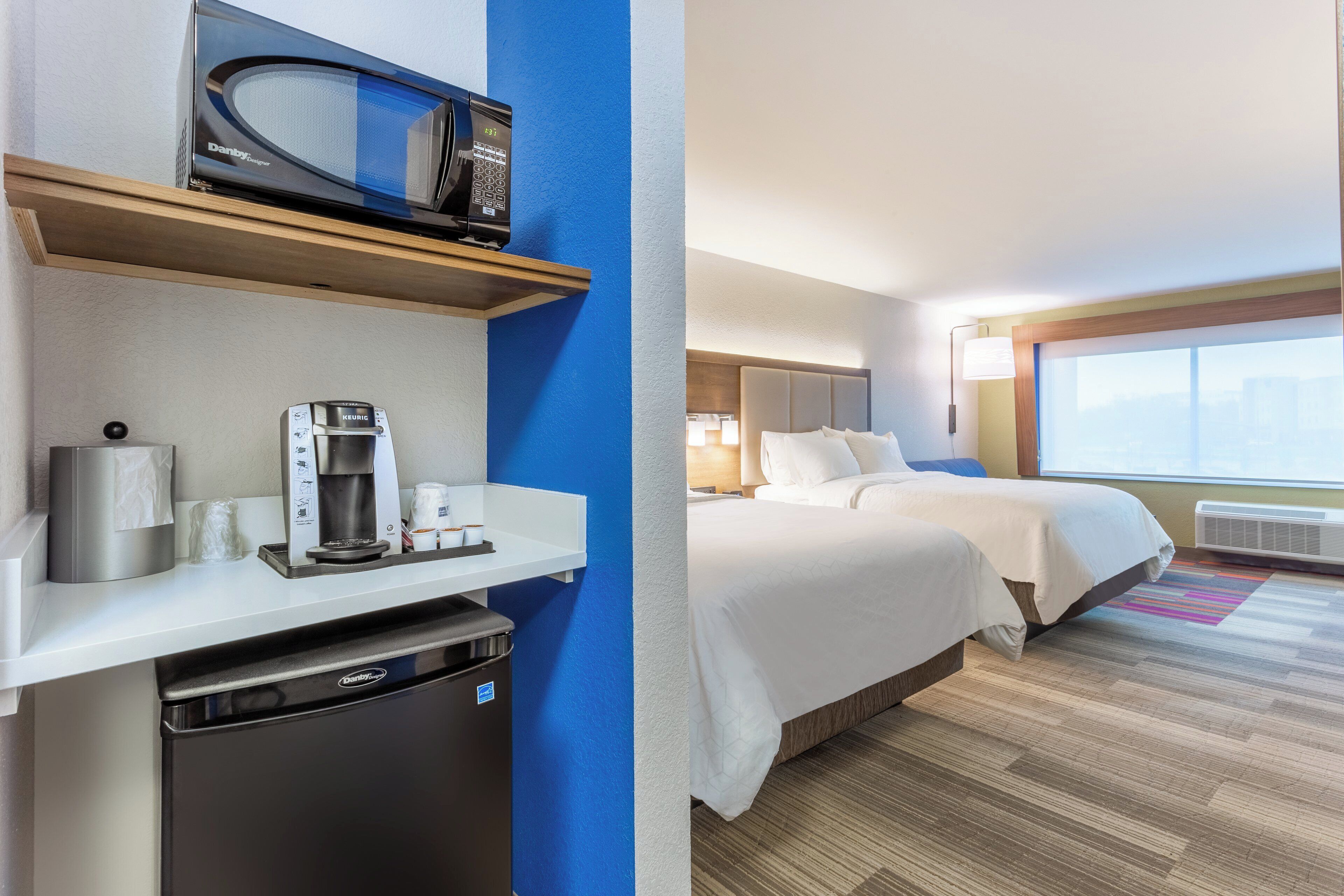 A hotel room with a kitchenette, a bed, a window with a city view, and a bathroom.