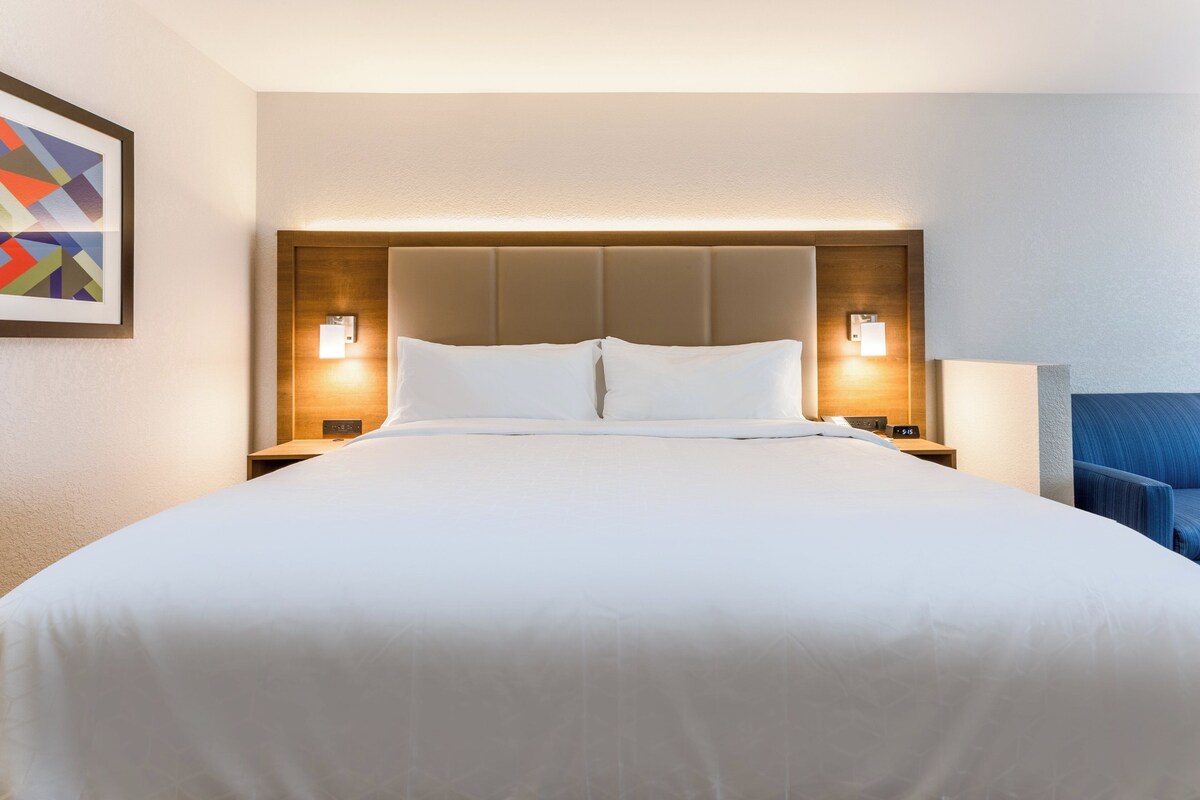 Habitación de hotel con una cama grande, dos mesitas de noche con lámparas, una silla azul y un cuadro enmarcado en la pared.