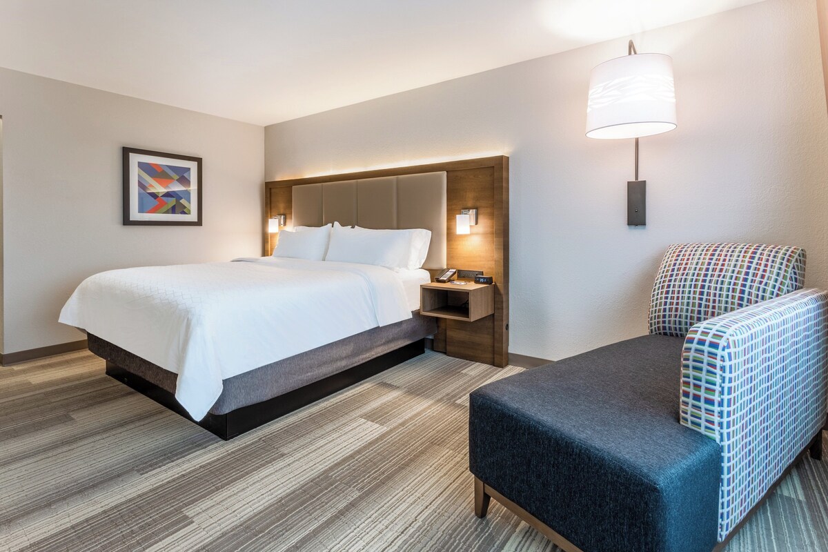 Habitación de hotel con cama, mesita de noche, una silla, una lámpara de pared y un cuadro en la pared.