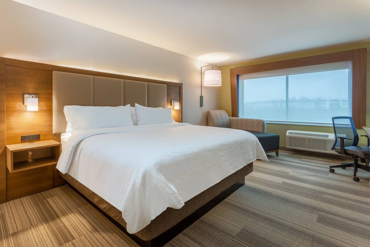 A hotel room with a large bed, bedside tables, a chair, a window, and a view of the city.