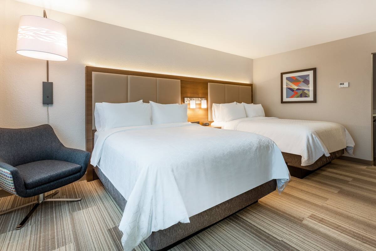 A hotel room with two beds, a chair, a lamp, and a painting on the wall.