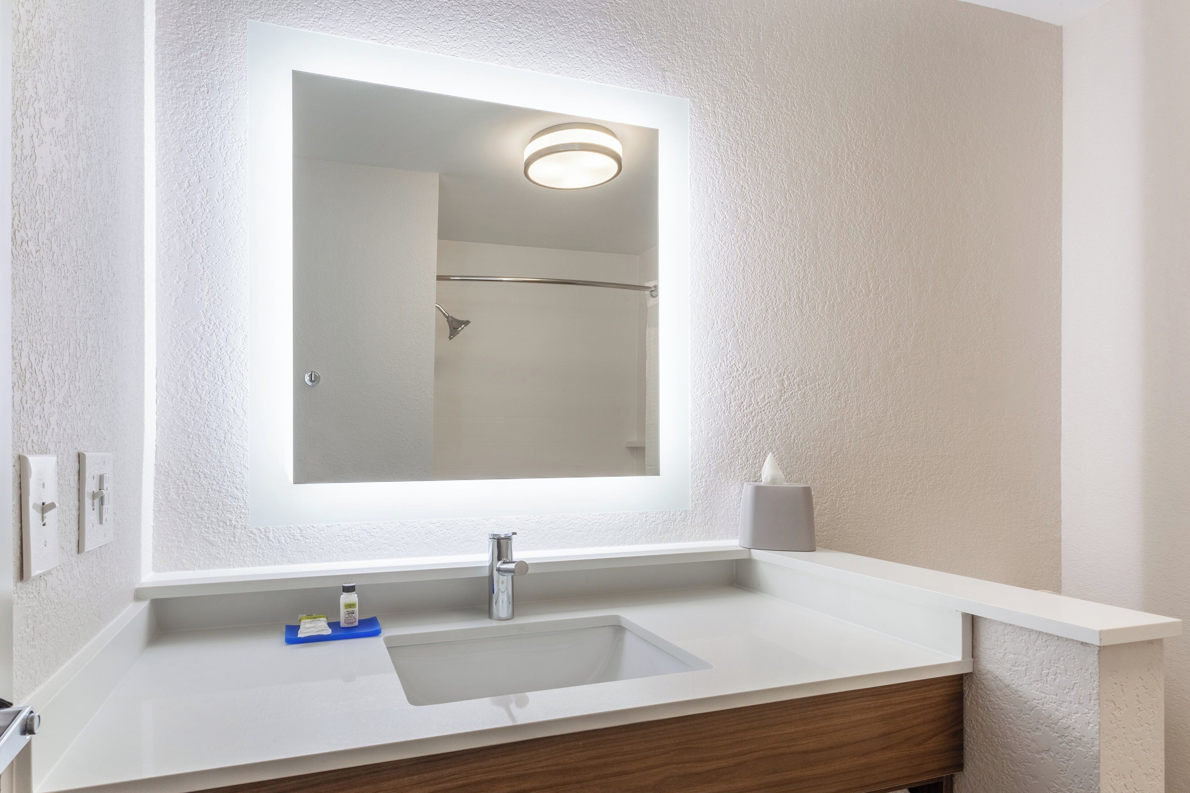 A bathroom with a white sink, a mirror with a light, a towel dispenser, and a shower area.