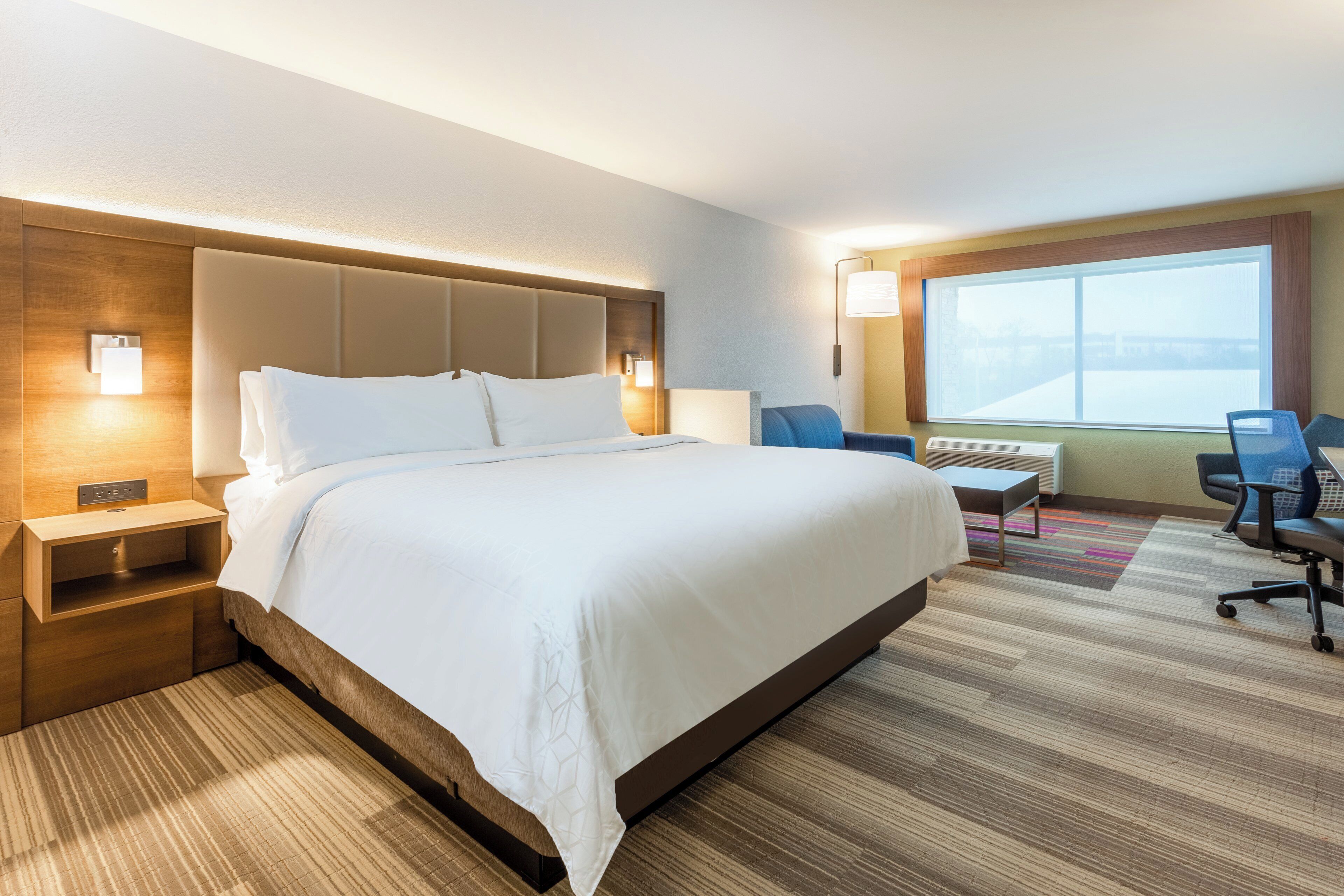 Une chambre d’hôtel avec un grand lit, des tables de chevet, un bureau, une chaise et une grande fenêtre.