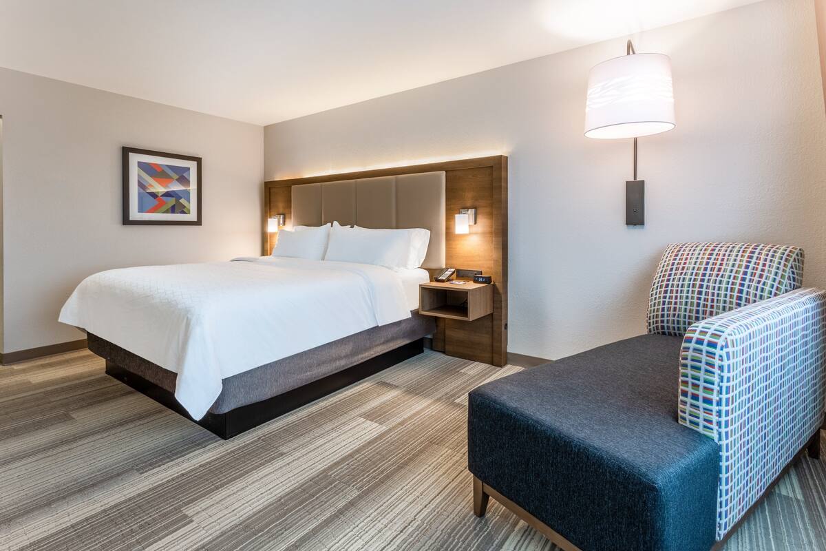 A hotel room with a bed, a nightstand, a chair, a wall-mounted lamp, and a framed picture on the wall.