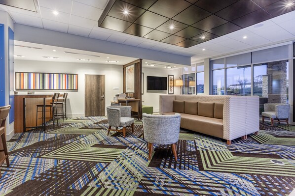 Un moderno vestíbulo de hotel con alfombra estampada, un sofá, sillas, zona de bar y grandes ventanales.