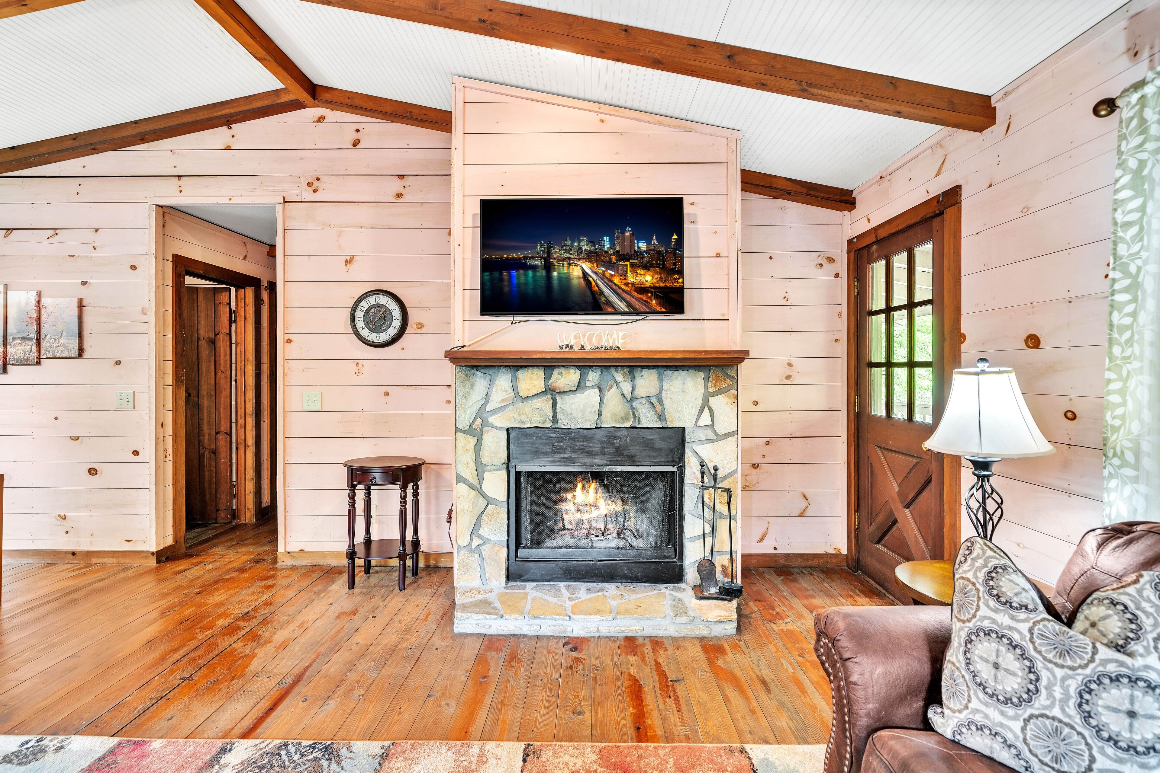A cozy living room with a stone fireplace, wooden floors, a television mounted above the fireplace, and a leather sofa.