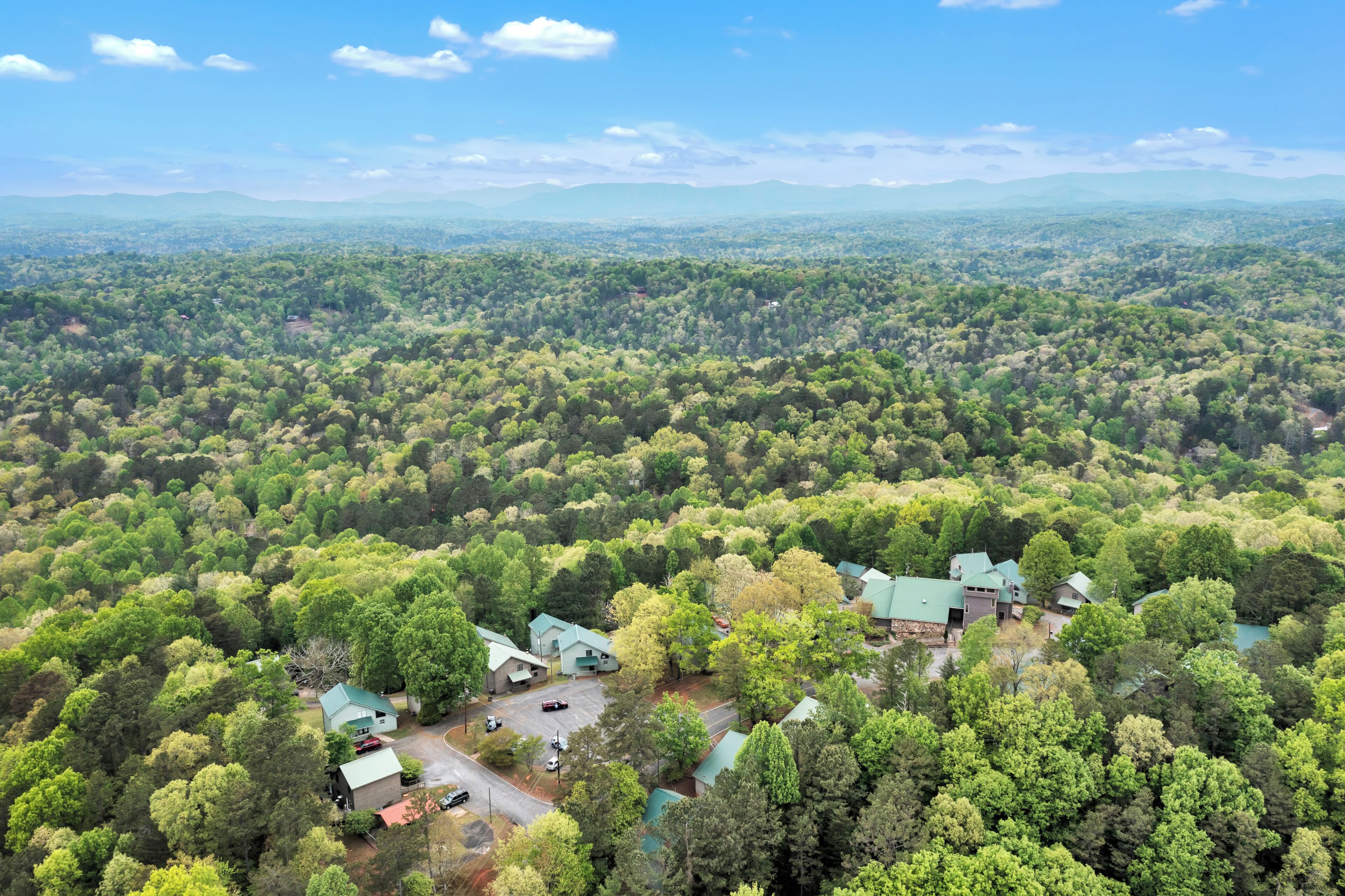 An aerial view of a forested area with a cluster of buildings featuring green roofs, surrounded by dense trees and a distant mountain range.
