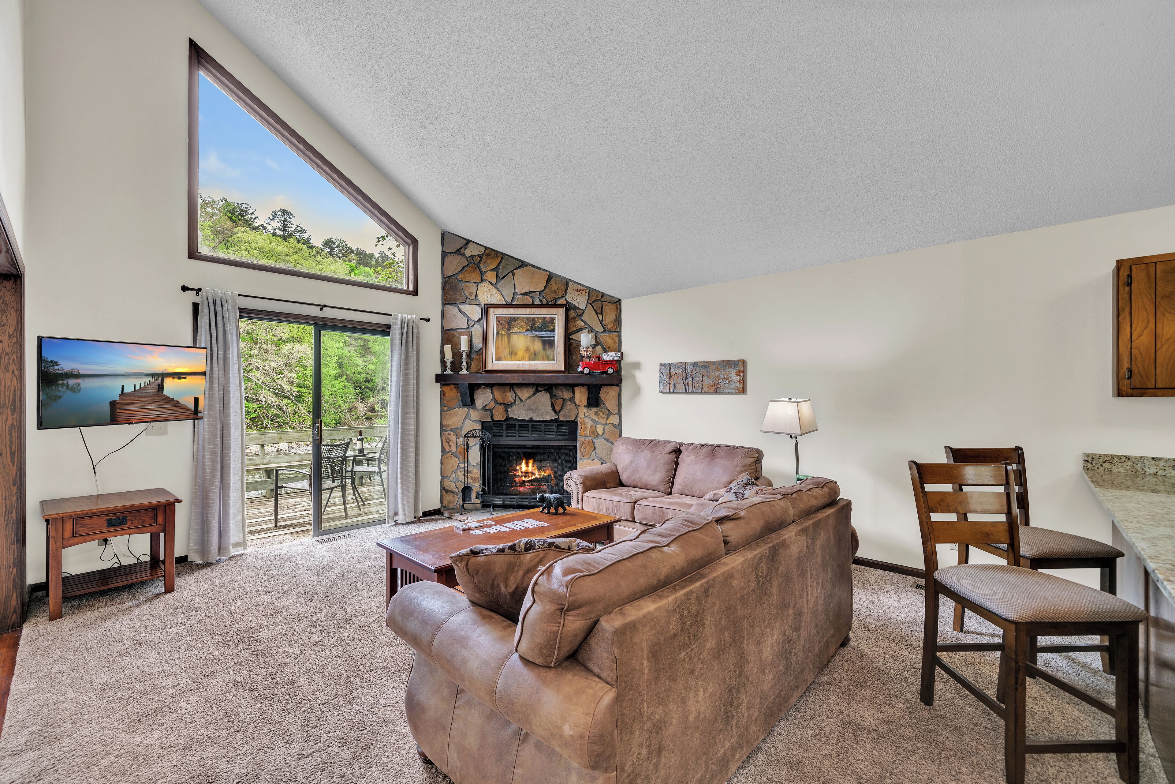 A living room with a stone fireplace, leather sofa, and a large window overlooking a deck.