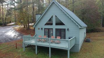 A small, light blue cabin with a porch and two chairs, surrounded by trees and a parking area.