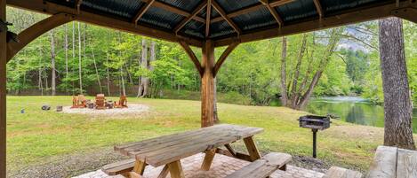 A covered picnic area with wooden benches and tables, surrounded by a grassy field and a pond.