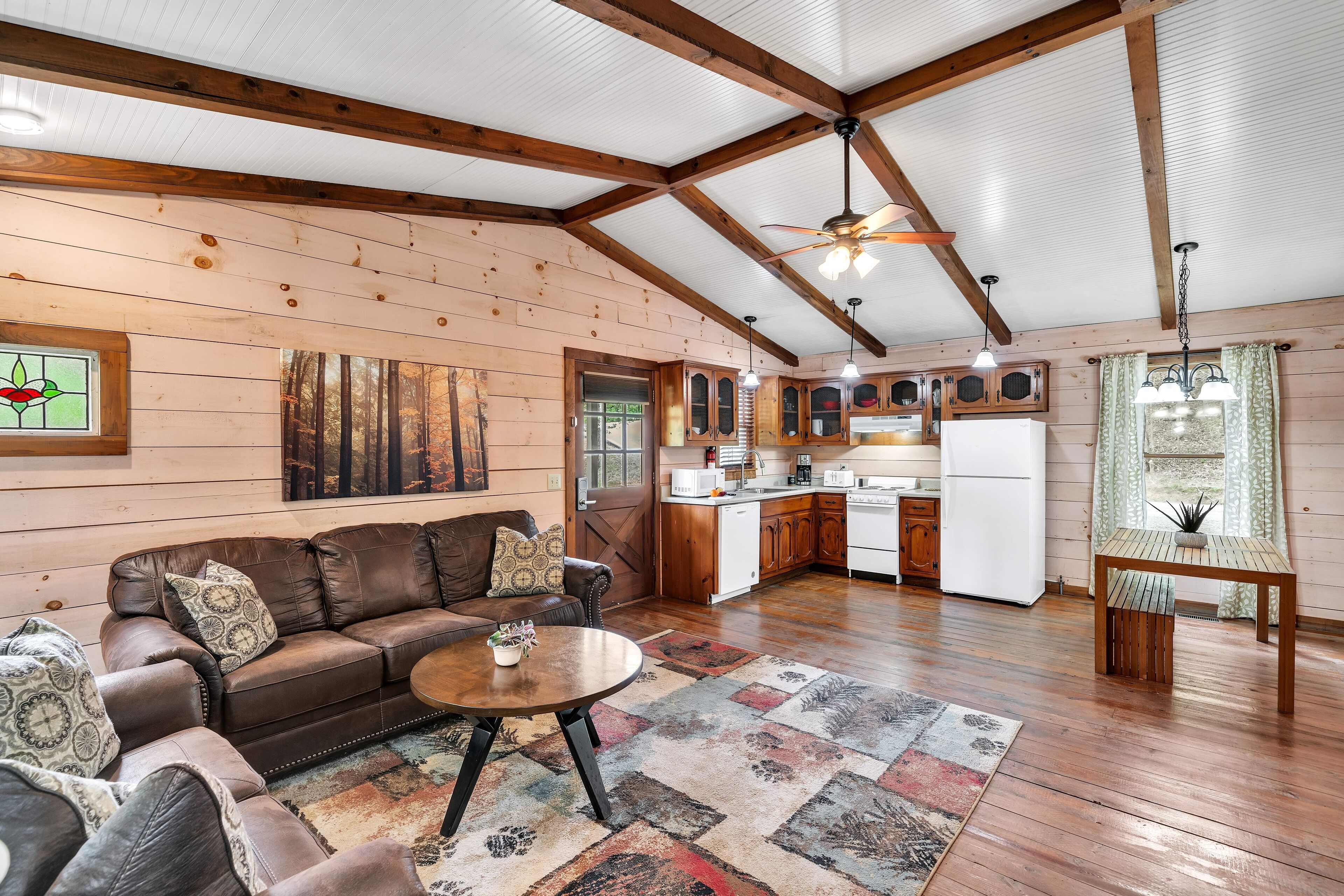 A cozy cabin interior with a wooden ceiling, a living room with a brown leather sofa, a dining table, and a kitchen area.
