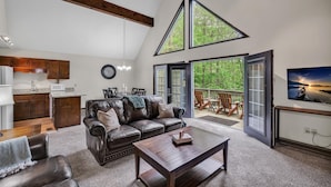 A spacious living area with a high ceiling, a dark leather sofa, a wooden coffee table, and a large window offering a view of a forest.