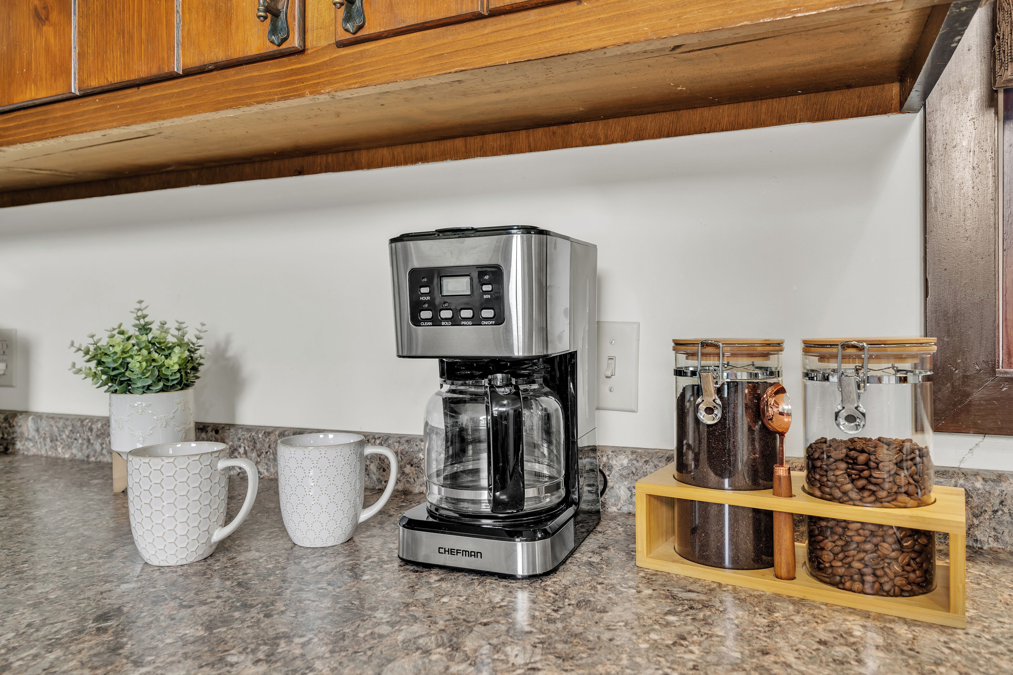 Een keuken met een koffiezetapparaat, twee mokken, een potplant en koffiebonen in potten.