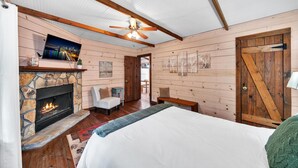 A cozy bedroom with a stone fireplace, a bed, a wooden ceiling fan, and a view of the city at night.
