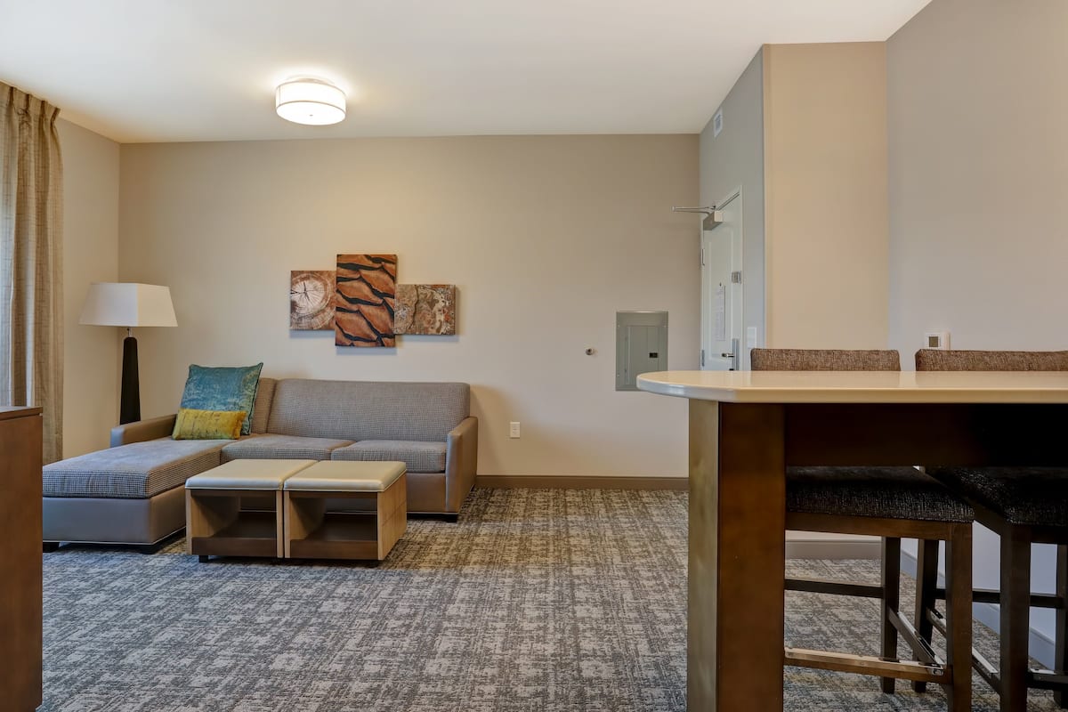 A compact living space with a sofa, ottoman, and a small table. There are two wall-mounted artworks and a ceiling light.