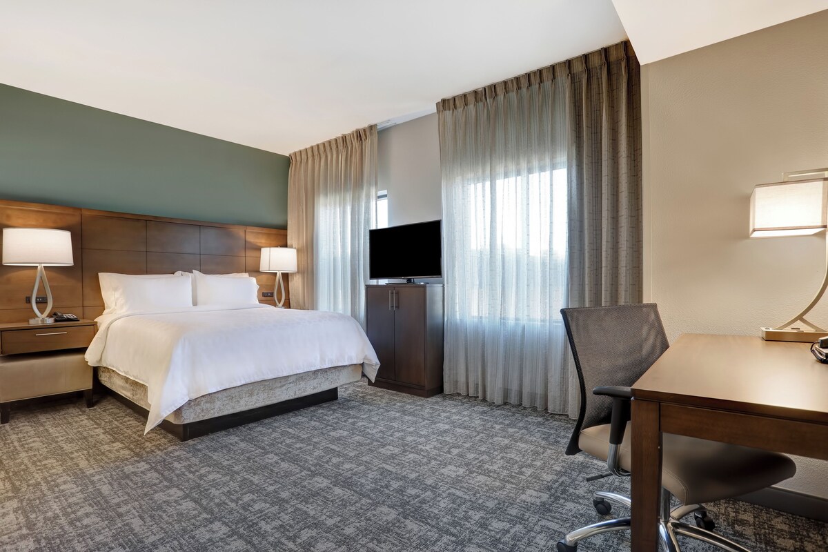 Habitación de hotel con cama, lámparas de noche, escritorio con silla, televisor y una ventana grande con cortinas.