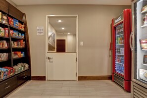 Un pasillo con una máquina expendedora, estantes con aperitivos y una puerta que da a otra habitación.