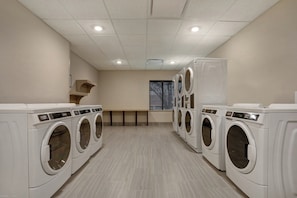 A laundry room with multiple white front-loading washing machines, a bench, and a window with blinds. - Staybridge Suites Overland Park - Kansas City S by IHG (Overland Park)