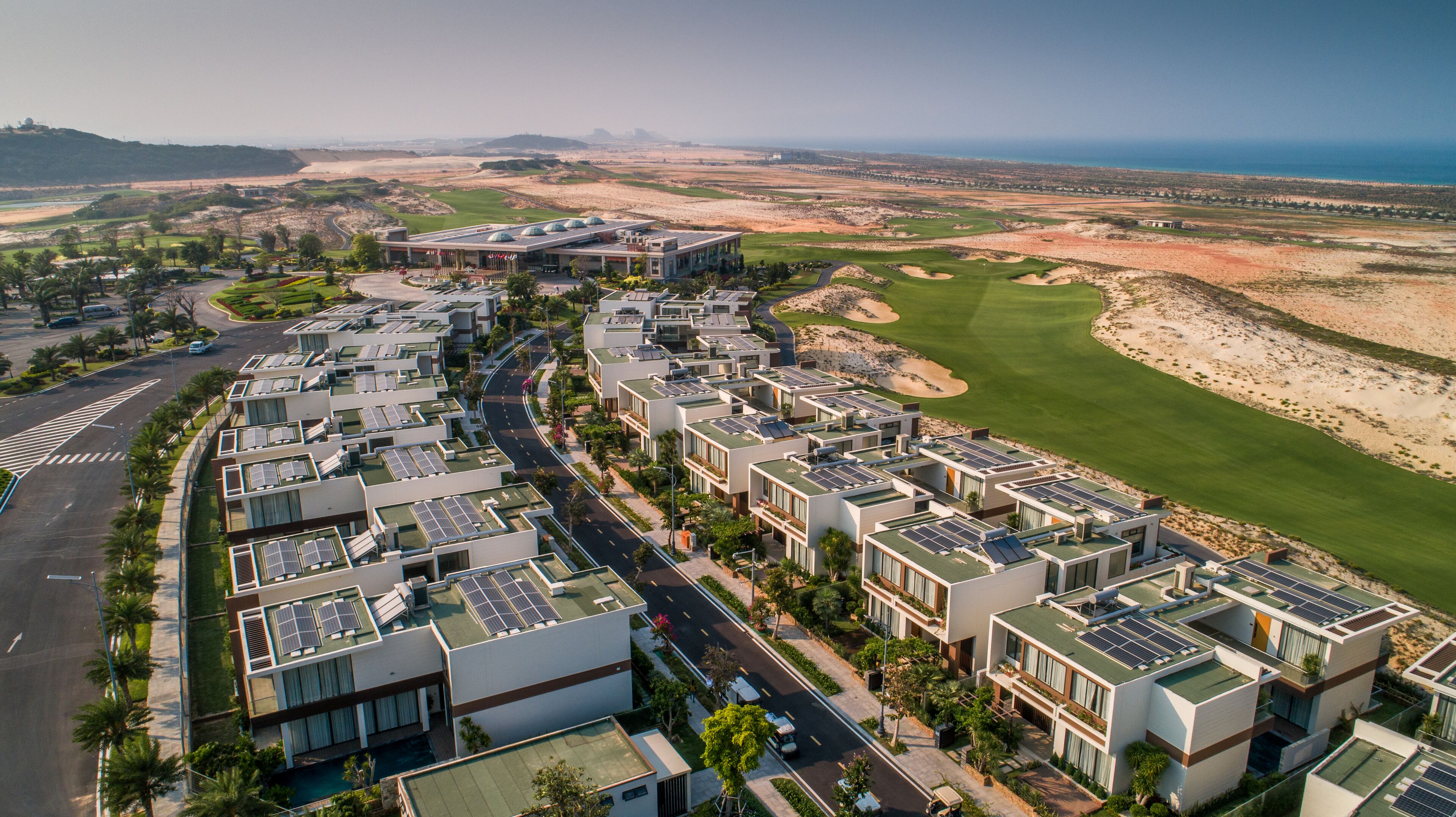 A modern residential area with solar panels on the rooftops, a golf course nearby, and a clear view of the coastline.