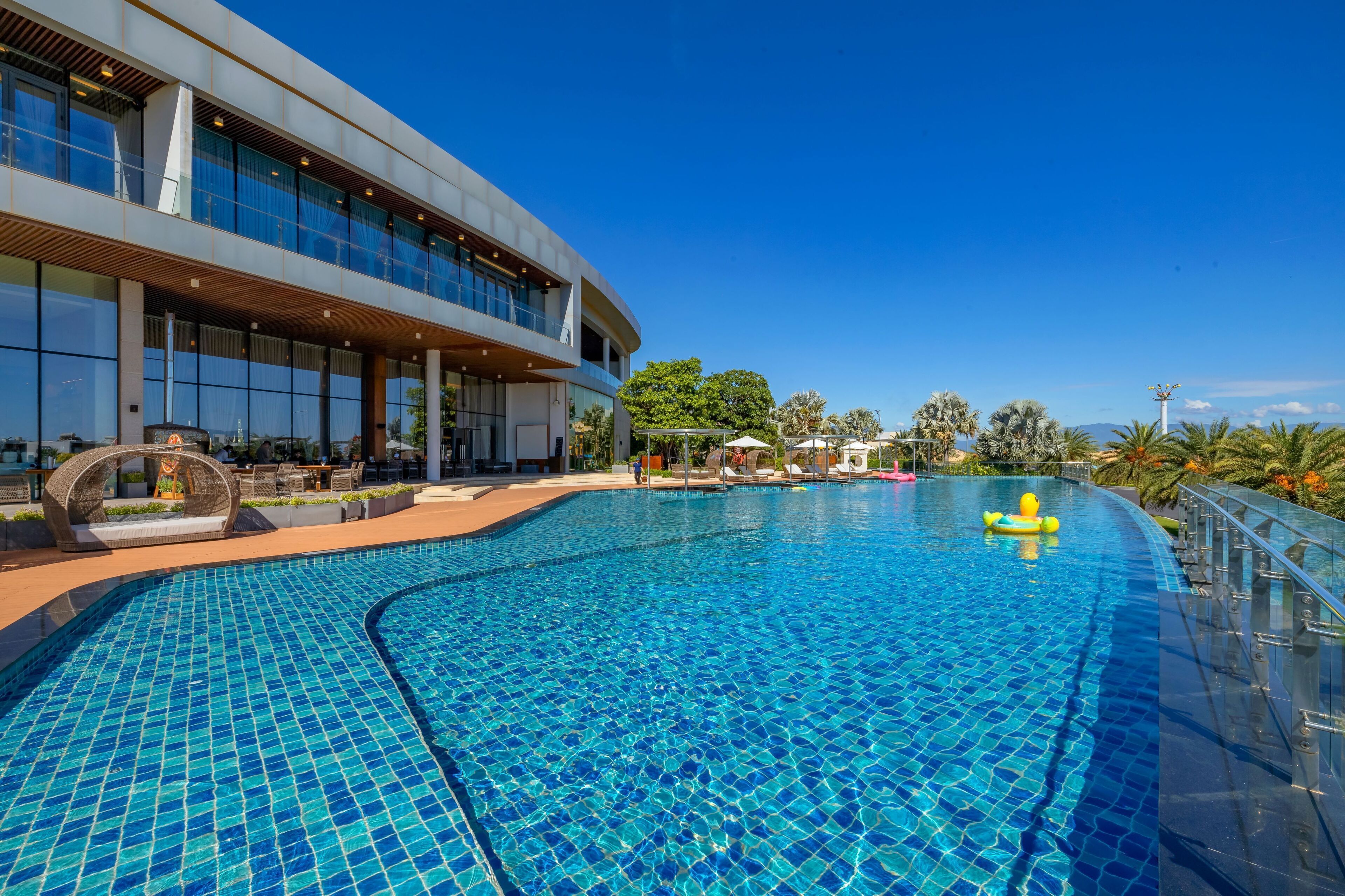 A modern hotel with a large swimming pool, outdoor seating, and a clear blue sky.