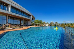 A modern hotel with a large swimming pool, outdoor seating, and a clear blue sky.