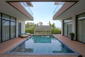 A modern outdoor swimming pool with a stone wall and wooden deck, surrounded by glass doors and windows. - Wyndham Grand KN Paradise Cam Ranh (Bac Cam Ranh)