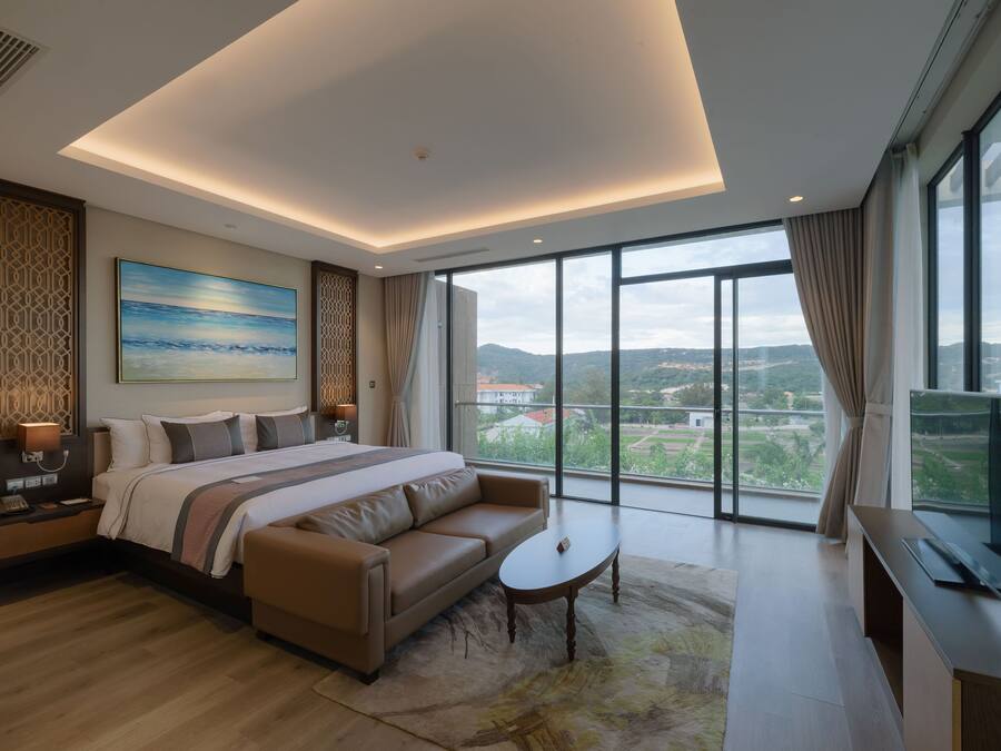 A modern hotel room with a large bed, a sofa, a small table, and a TV. The room has large windows offering a view of the city.
