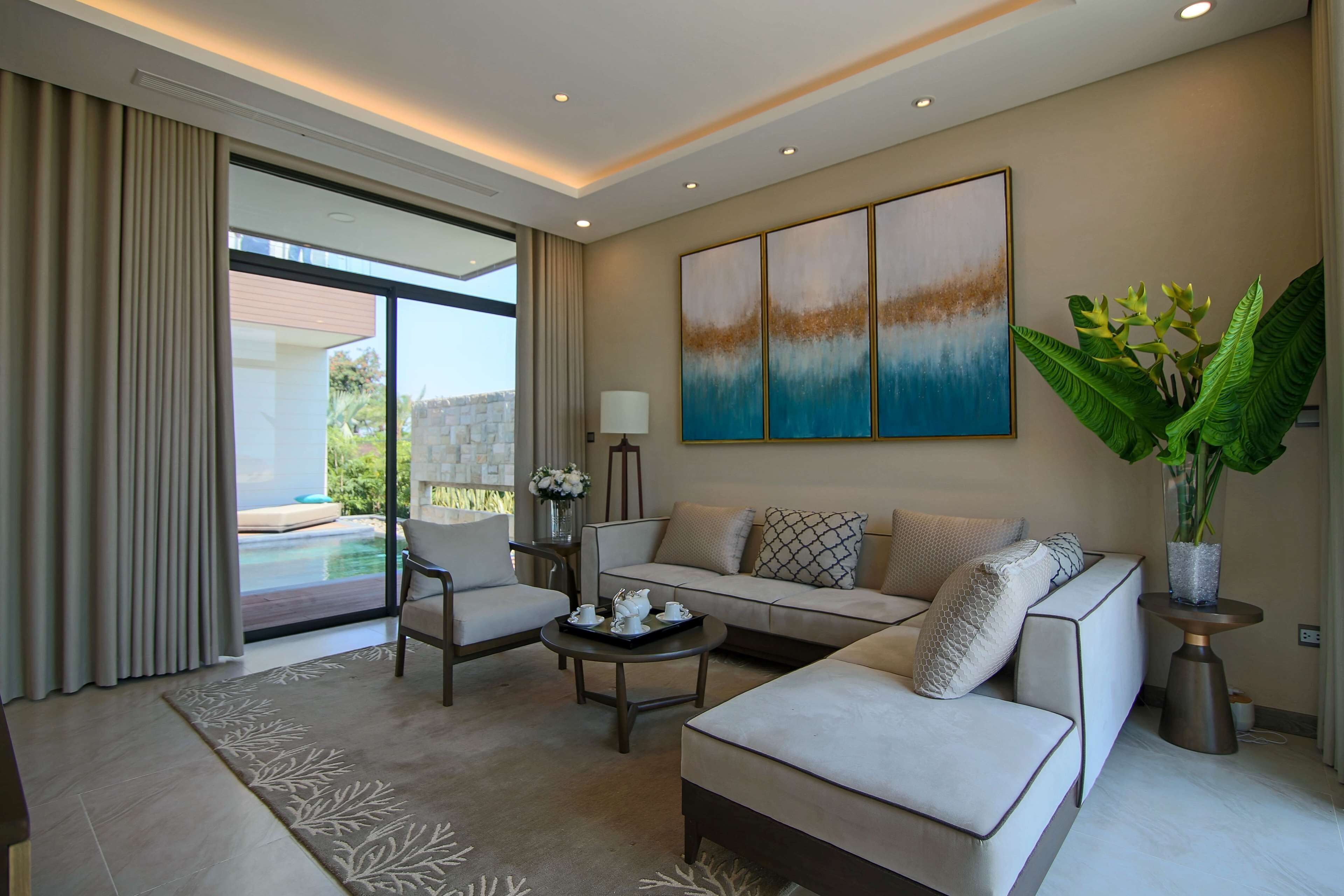 A modern living room with a sofa, armchair, and coffee table. There is a large window with curtains, a painting on the wall, and a potted plant.