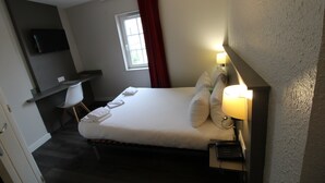 Une chambre d’hôtel avec un lit, une lampe de chevet, un bureau avec une chaise, un téléviseur fixé au mur et une fenêtre avec des rideaux rouges.