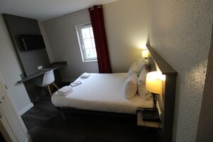 Una habitación de hotel con cama, lámpara de noche, escritorio con silla, televisor montado en la pared y ventana con cortinas rojas.