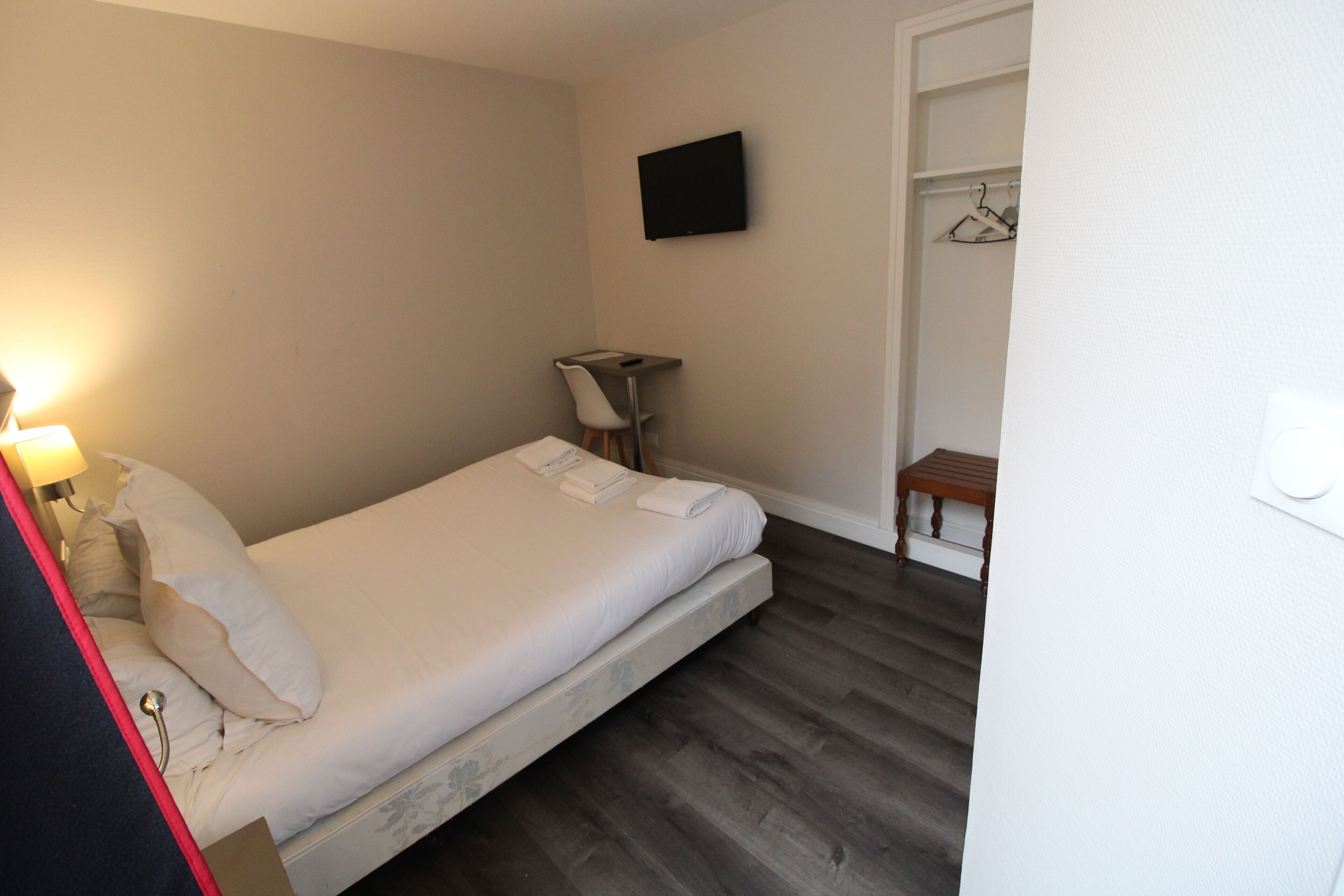 Een kleine, verzorgde slaapkamer met een enkel bed, een nachtkastje, een stoel, een aan de muur bevestigde televisie en een houten bankje.