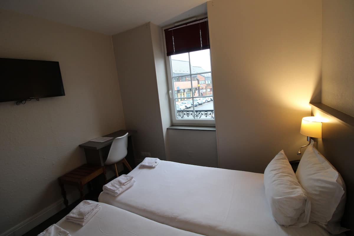 Hotelkamer met twee bedden, een bureau, een stoel, een aan de muur bevestigde televisie en een raam met uitzicht op gebouwen en geparkeerde auto's.