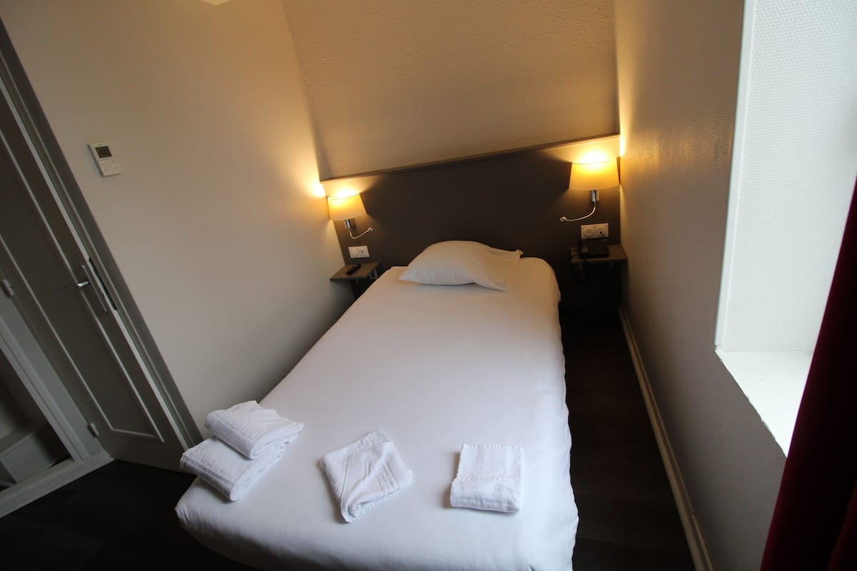 Een keurig opgemaakt bed met witte lakens en handdoeken, twee bedlampjes, en een deur links.