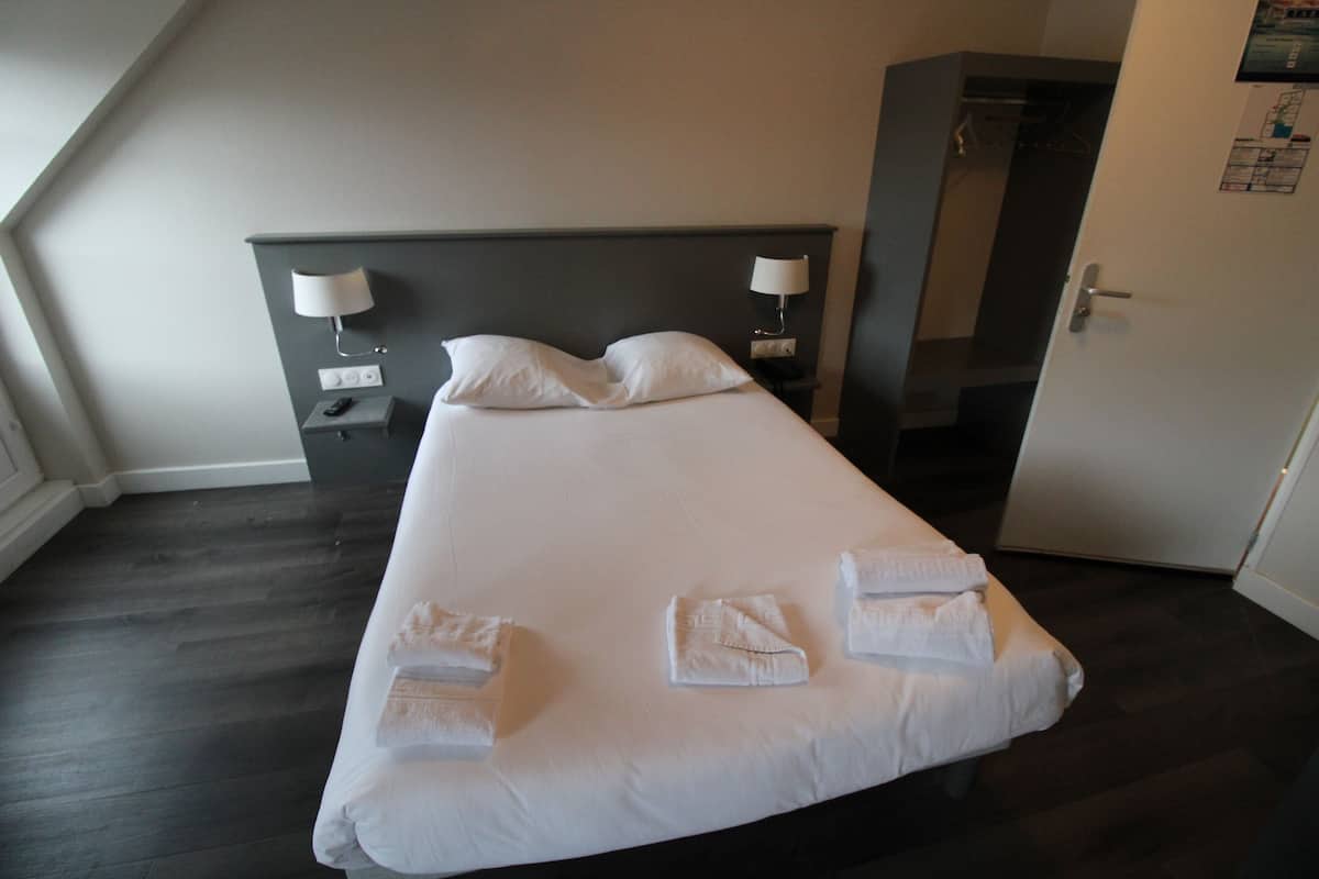 Een netjes opgemaakt bed met witte lakens en handdoeken, een donker houten vloer, een hoofdbord met wandlampen en een deur met een kaart en informatie op de muur.