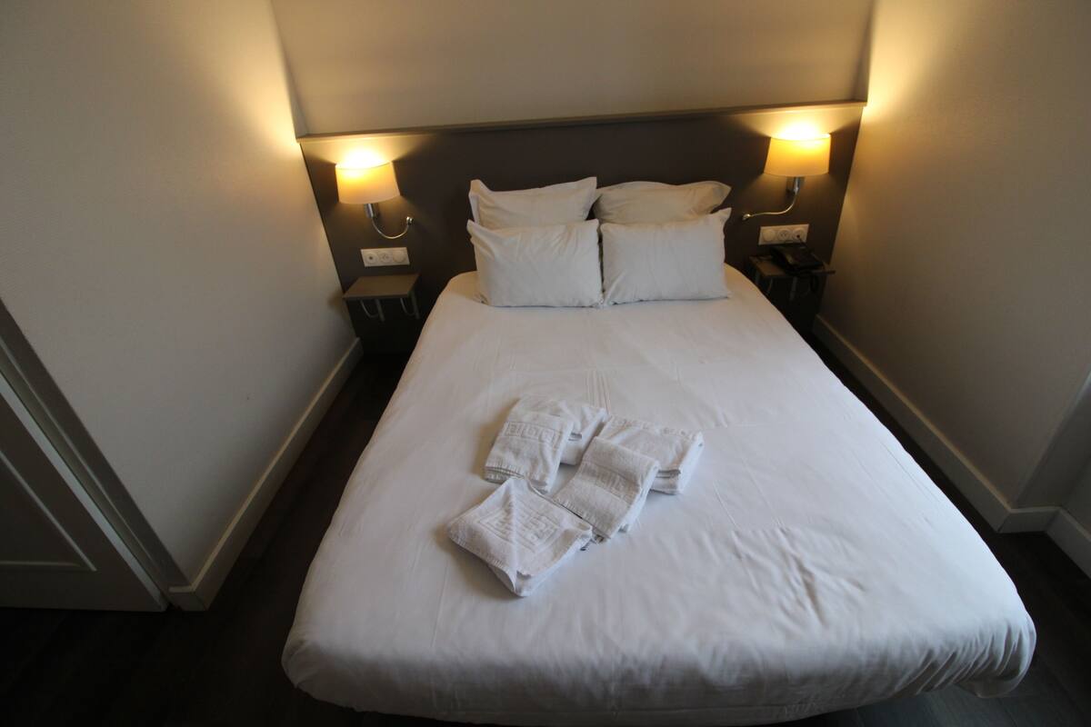 Een netjes opgemaakt bed met witte lakens en handdoeken, twee bedlampjes en een donker hoofdbord in een hotelkamer.