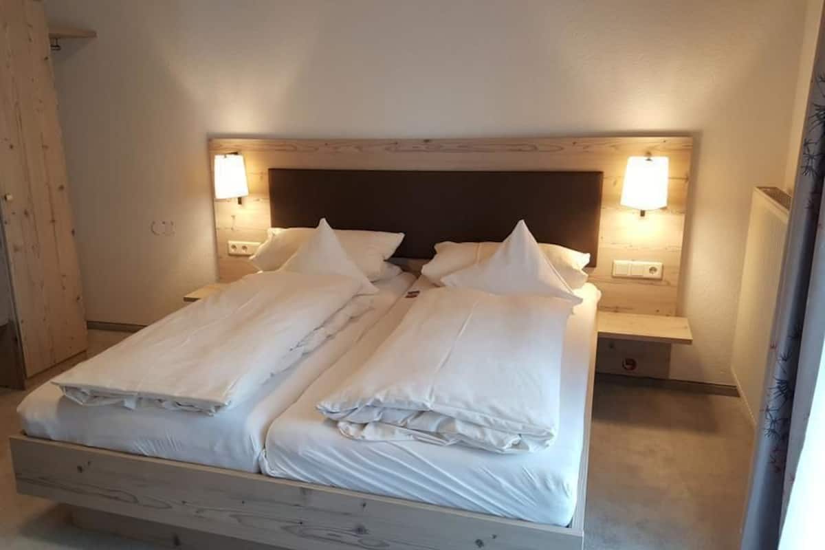 Un lit double avec du linge de lit blanc, une tête de lit en bois, deux tables de chevet avec des lampes et un banc en bois.
